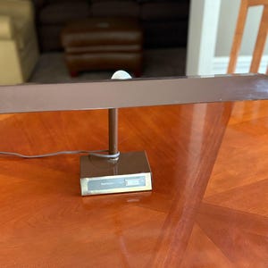 May include: A brown desk lamp with a rectangular shade and a square base. The lamp has a slender, vertical support and a small, white, spherical light bulb. The lamp is on a wooden table.