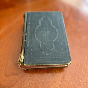 May include: A vintage, dark teal book with an embossed design on the cover. The cover features a cross within an ornate frame. The book's edges show signs of wear, with the spine partially detached. The book is resting on a wooden surface.