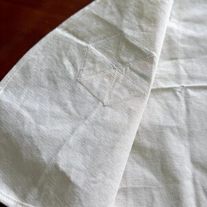 K&ouml;nnte beinhalten: Off-white Leinen-Serviette mit geometrischem Stickdesign. Die Serviette ist gefaltet und zeigt die gestickten Details an der Ecke. Der Stoff hat ein strukturiertes Aussehen, und die N&auml;hte sind sichtbar.