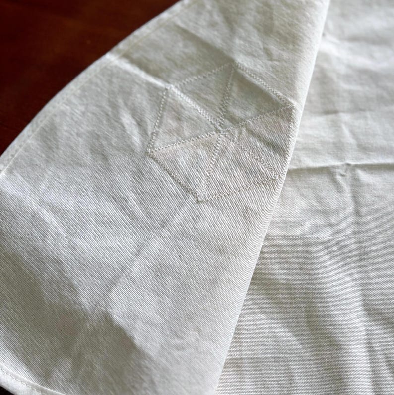 K&ouml;nnte beinhalten: Off-white Leinen-Serviette mit geometrischem Stickdesign. Die Serviette ist gefaltet und zeigt die gestickten Details an der Ecke. Der Stoff hat ein strukturiertes Aussehen, und die N&auml;hte sind sichtbar.