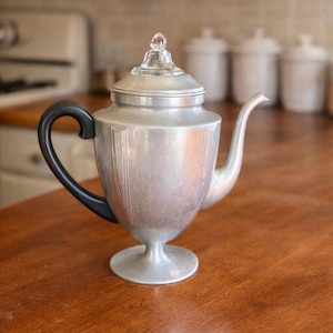 May include: A vintage silver-colored teapot with a black handle and a clear glass knob on the lid. The teapot has a curved spout and is set on a wooden surface. The design includes vertical lines on the body.