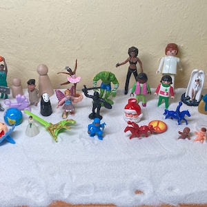 May include: A collection of colorful plastic figurines, including a ballerina, a Santa Claus, a frog, a fairy, a robot, a snowman, a dragon, a knight, a princess, a ghost, a tree, a bird, a flower, a person, and a cat. The figurines are arranged on a white surface.