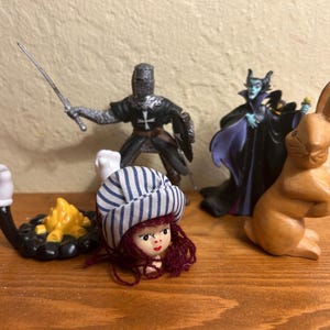 May include: A collection of figurines including a knight in armor with a sword, a witch in a black and purple gown, a wooden rabbit, a doll head with a striped hat, and a campfire.