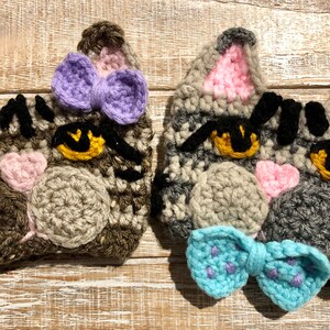 May include: Two crocheted cat faces with black eyes, pink noses, and gray and brown fur. One cat has a purple bow and the other has a blue bow.
