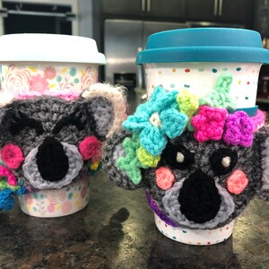 Puede incluir: Dos tazas de caf&eacute; con calientavasos de koala de ganchillo. Los calientavasos son grises con narices negras y mejillas rosadas. Una taza tiene un patr&oacute;n floral y la otra tiene lunares blancos. Ambos calientavasos tienen flores de colores.