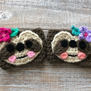 May include: Two crocheted sloth mug cozies with brown fur, white faces, pink cheeks, and black eyes. One has a pink flower and green leaves, the other has a purple flower and green leaves.
