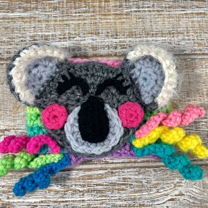 Puede incluir: Un koala de crochet con una cola de arco&iacute;ris. El koala es gris con ojos negros y mejillas rosadas. La cola est&aacute; hecha de hilo de colores.