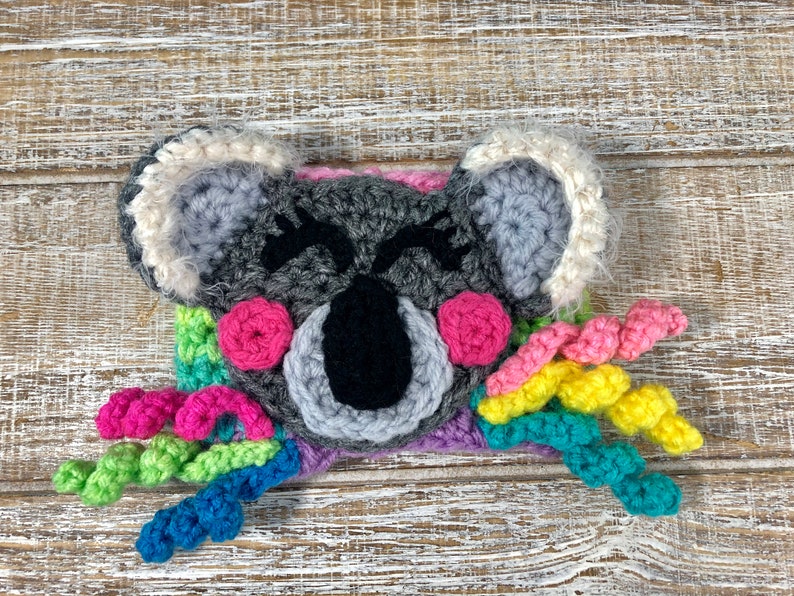 Puede incluir: Un koala de crochet con una cola de arco&iacute;ris. El koala es gris con ojos negros y mejillas rosadas. La cola est&aacute; hecha de hilo de colores.