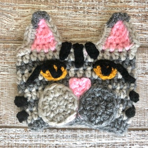 May include: Crocheted gray cat face with black stripes, pink ears, and yellow eyes. The cat has a pink heart nose and gray crocheted whiskers.