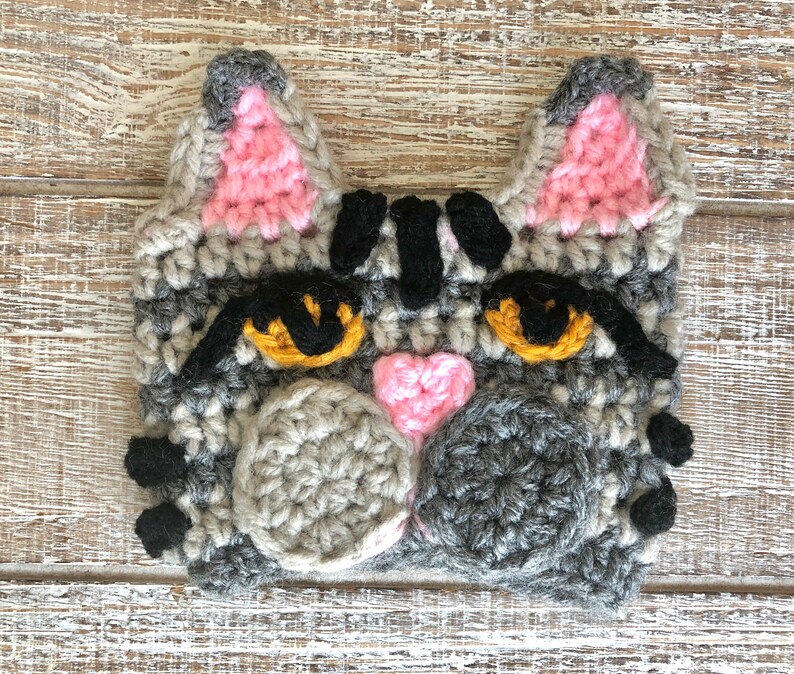 May include: Crocheted gray cat face with black stripes, pink ears, and yellow eyes. The cat has a pink heart nose and gray crocheted whiskers.