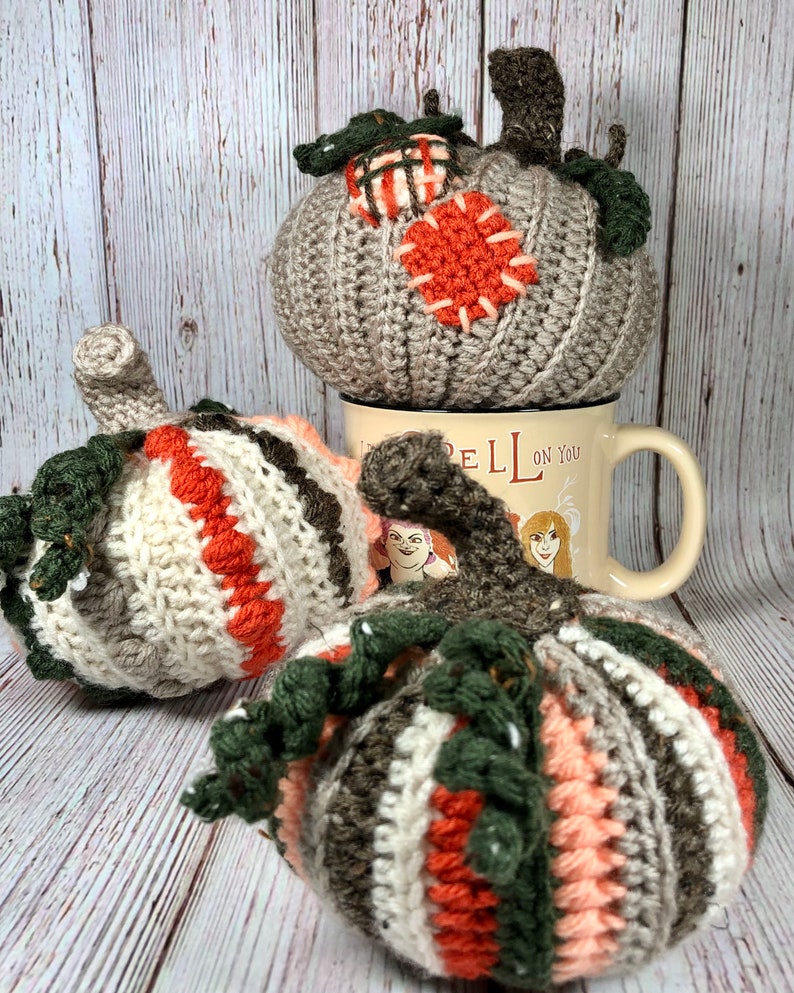 May include: Handmade crochet pumpkins in various colors, including gray, orange, white, and brown. One pumpkin rests on a mug with the text "Spell on You". The pumpkins have green stems and decorative patches.