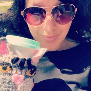 May include: A woman wearing sunglasses holds a crocheted cat-shaped coffee cup cozy. The cozy is gray and white with pink accents and black eyes. The cup is white with a light blue lid.