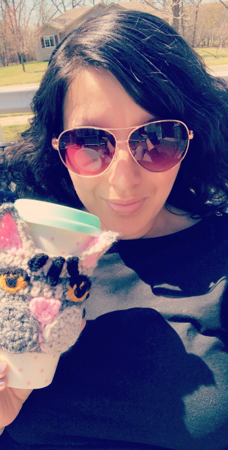 May include: A woman wearing sunglasses holds a crocheted cat-shaped coffee cup cozy. The cozy is gray and white with pink accents and black eyes. The cup is white with a light blue lid.