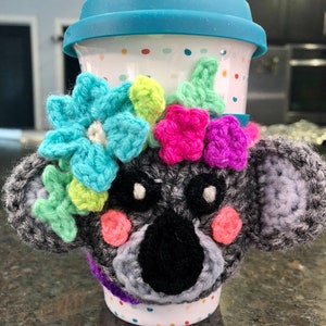 Puede incluir: Un calentador de taza de koala de crochet gris con flores de crochet de colores alrededor de la parte superior. El calentador est&aacute; en una taza de viaje de cer&aacute;mica blanca con lunares de colores.