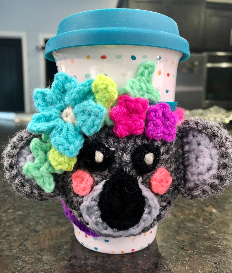 Puede incluir: Un calentador de taza de koala de crochet gris con flores de crochet de colores alrededor de la parte superior. El calentador est&aacute; en una taza de viaje de cer&aacute;mica blanca con lunares de colores.