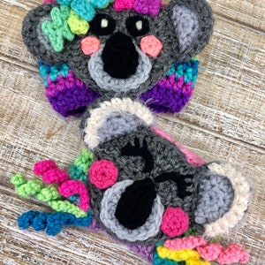 Puede incluir: Dos gorros de koala tejidos a crochet con flores y remolinos coloridos. Los gorros son grises con ojos negros y mejillas rosadas.