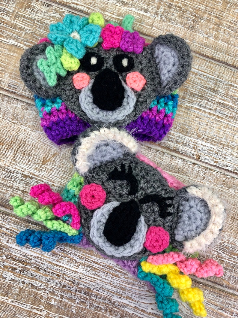 Puede incluir: Dos gorros de koala tejidos a crochet con flores y remolinos coloridos. Los gorros son grises con ojos negros y mejillas rosadas.