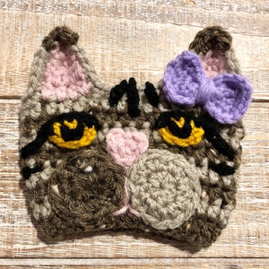 May include: A crocheted cat face with brown and gray yarn, black eyes, a pink nose, and a purple bow.