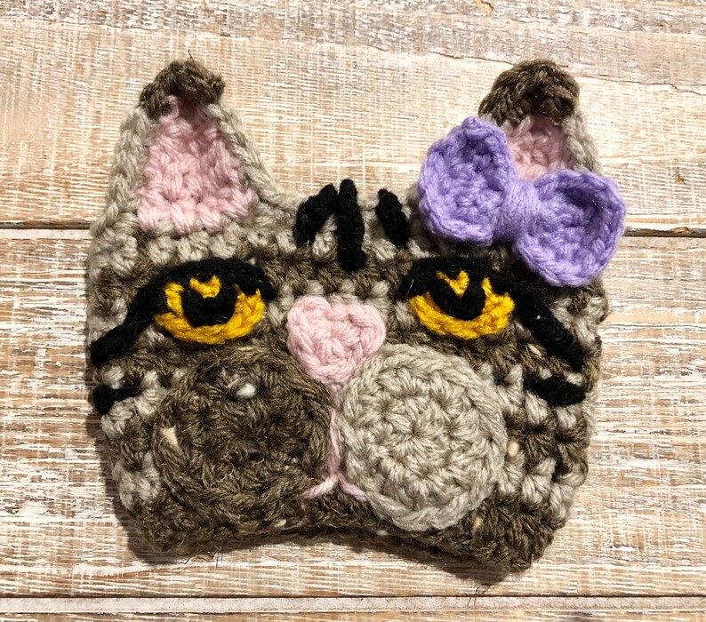 May include: A crocheted cat face with brown and gray yarn, black eyes, a pink nose, and a purple bow.