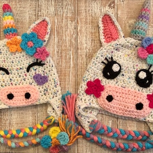 May include: Two white crocheted unicorn hats with pink ears, black eyes, and pink noses. Each hat has a colorful crocheted horn and a flower on top. The hats have long, colorful braided ties.