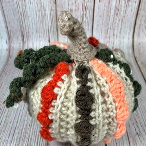 May include: A handmade crochet pumpkin in shades of cream, orange, brown, peach, and green. The pumpkin has a textured surface with a stem and leaves. This decorative item is perfect for fall and autumn decor.