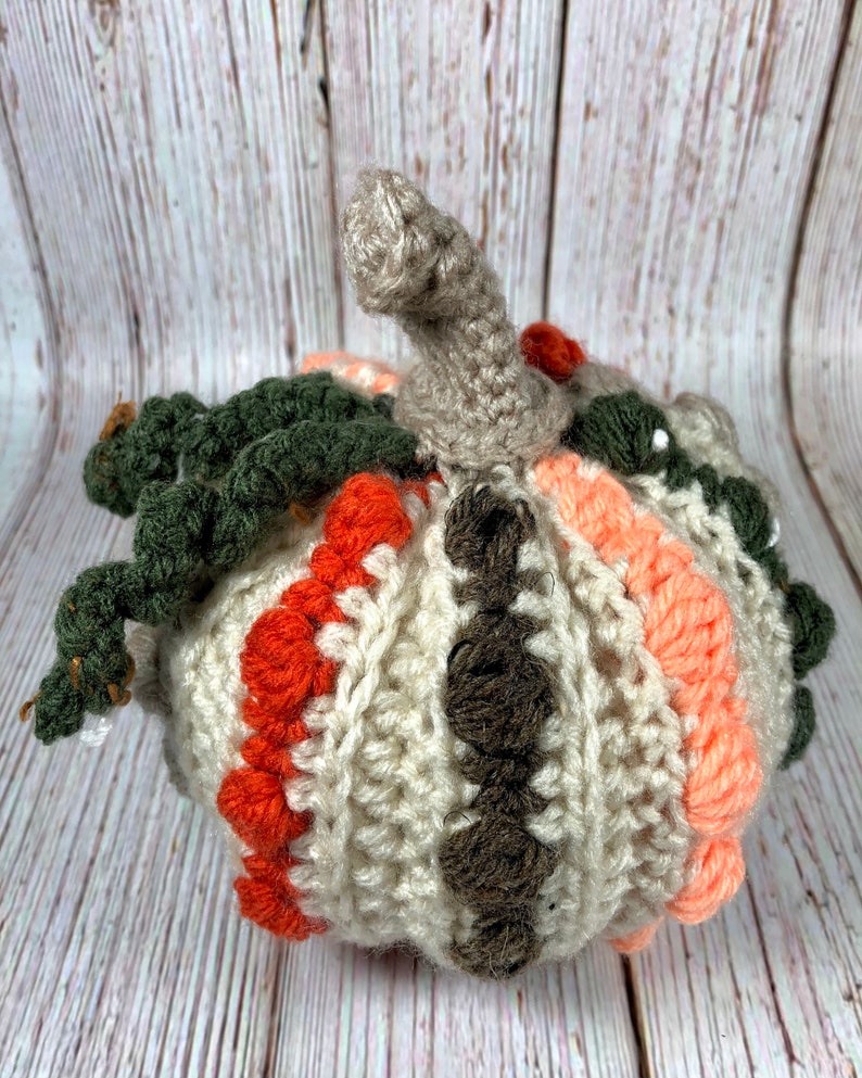 May include: A handmade crochet pumpkin in shades of cream, orange, brown, peach, and green. The pumpkin has a textured surface with a stem and leaves. This decorative item is perfect for fall and autumn decor.