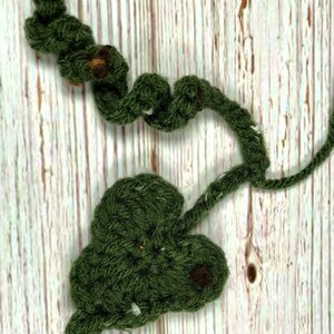 May include: Handmade crochet vine and leaf in a deep green color. The vine has a twisted shape with small brown accents, and the leaf is heart-shaped. The piece is set against a light wood grain background.