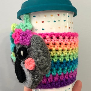 Puede incluir: Una taza de viaje de cer&aacute;mica blanca con una tapa azul verdosa y un acogedor de crochet arco&iacute;ris. El acogedor presenta una cara de animal de crochet gris con acentos rosados.