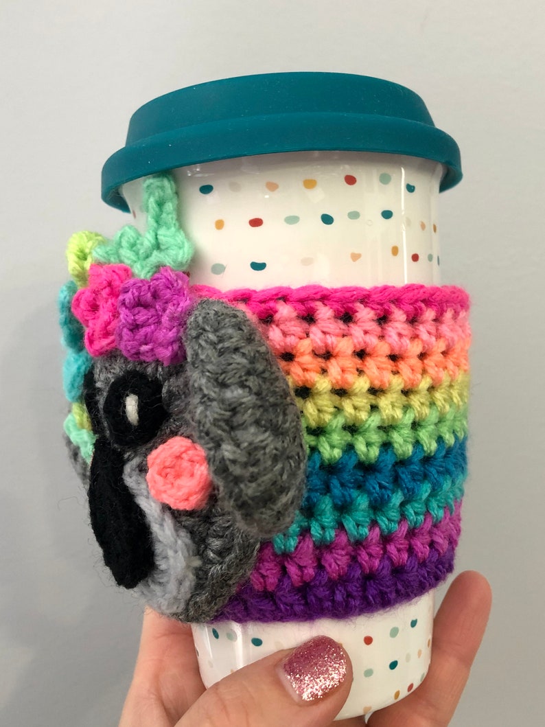 Puede incluir: Una taza de viaje de cer&aacute;mica blanca con una tapa azul verdosa y un acogedor de crochet arco&iacute;ris. El acogedor presenta una cara de animal de crochet gris con acentos rosados.
