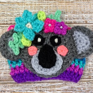 Puede incluir: Un gorro de koala de crochet gris con mejillas rosadas, ojos negros y nariz negra. El gorro est&aacute; decorado con flores de crochet de colores y lazos rosas con pedrer&iacute;a.