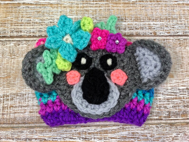 Puede incluir: Un gorro de koala de crochet gris con mejillas rosadas, ojos negros y nariz negra. El gorro est&aacute; decorado con flores de crochet de colores y lazos rosas con pedrer&iacute;a.