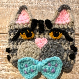 May include: Gray crocheted cat face with pink ears and a blue bow tie. The cat has black eyes and a black nose.