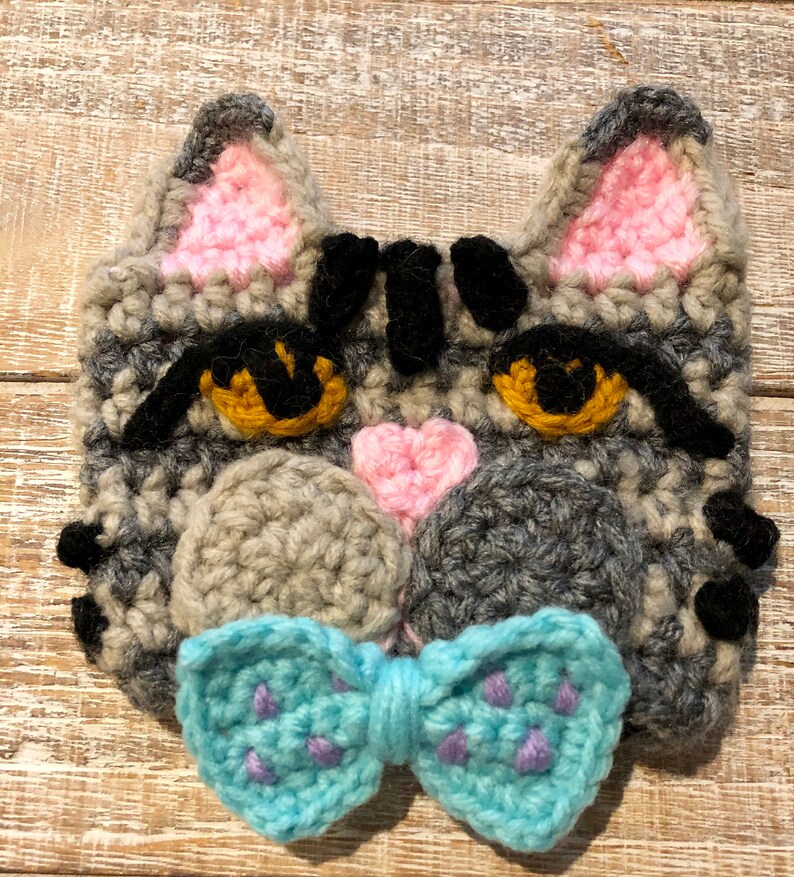 May include: Gray crocheted cat face with pink ears and a blue bow tie. The cat has black eyes and a black nose.