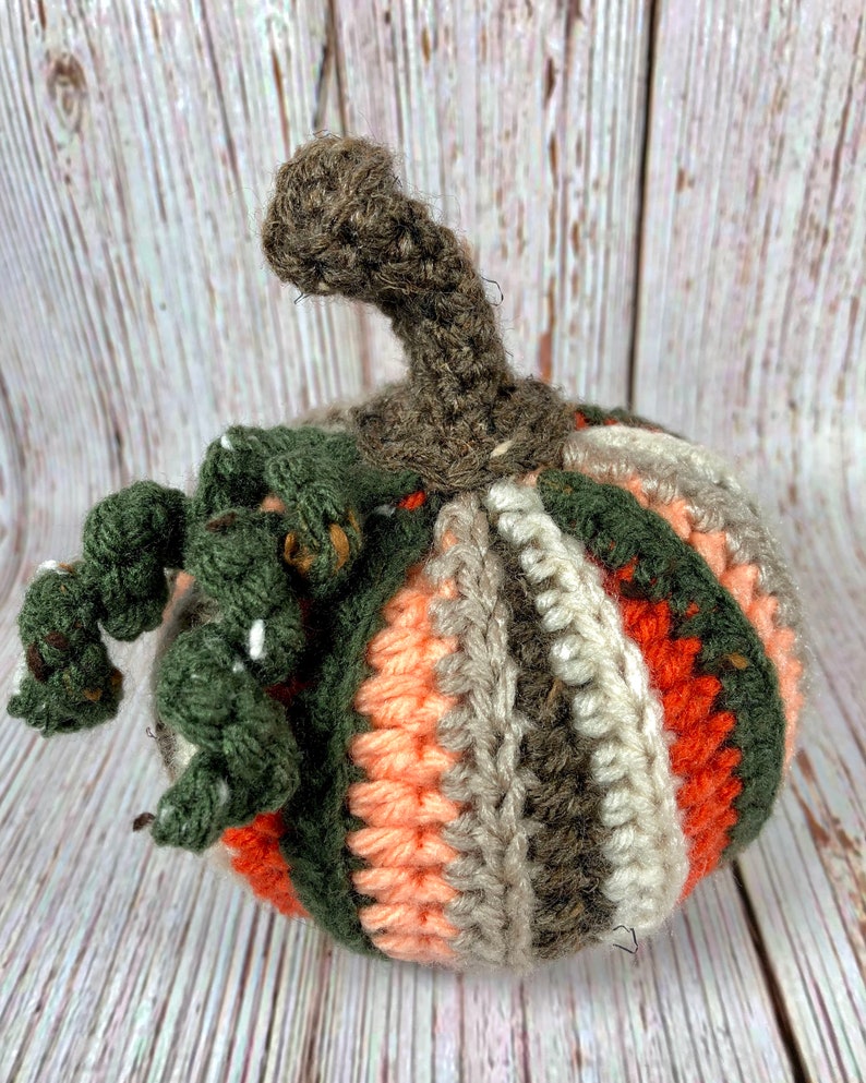 May include: A handmade crochet pumpkin in shades of orange, green, gray, and brown. The pumpkin has a stem and vine detail. The pumpkin is a decorative item.