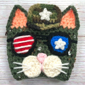 May include: A crocheted cat with a camouflage hat, red and white striped eyes, blue and white star eyes, and a white nose.
