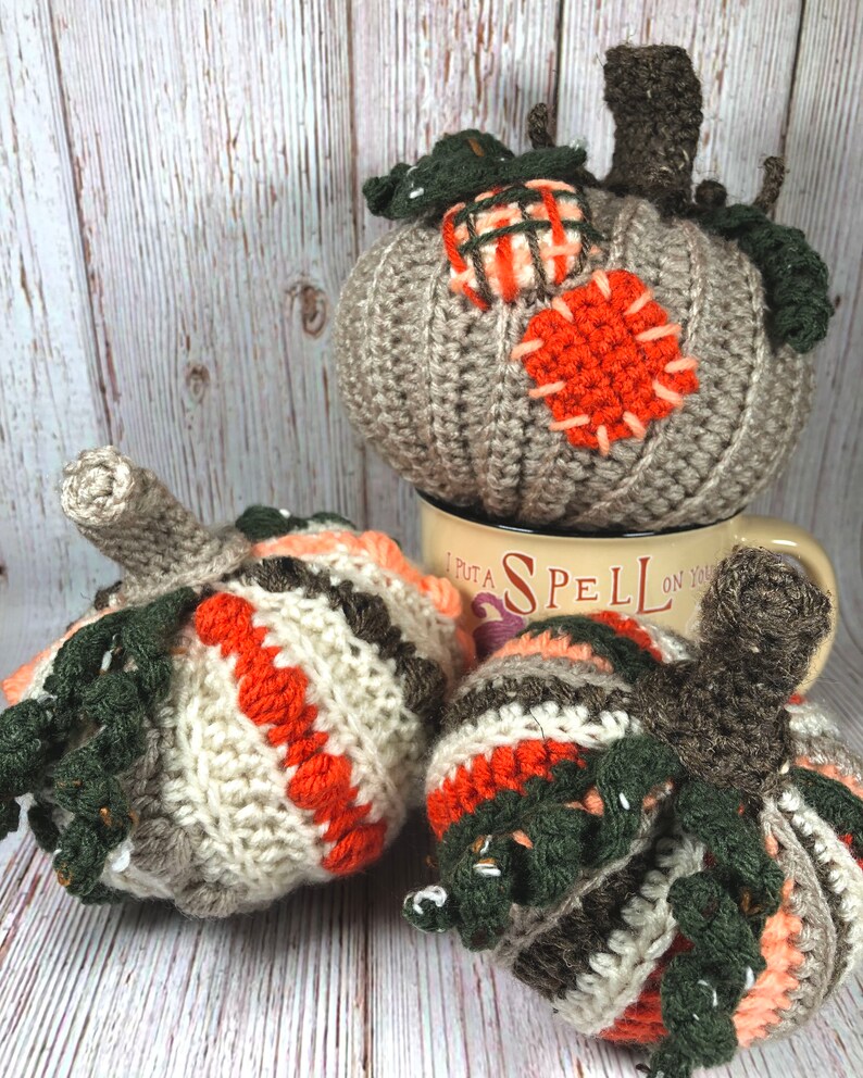 May include: A collection of handmade crochet pumpkins in various sizes and colors. The pumpkins feature shades of gray, orange, white, and green, with detailed stitching and stem accents. One pumpkin rests on a mug with the text "I PUT A SPELL ON YOU".