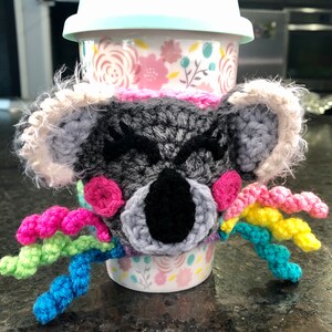 Puede incluir: Un calientavasos de ganchillo de koala con detalles de hilo arco&iacute;ris. El calientavasos es gris con detalles rosas y ojos negros. El calientavasos est&aacute; en una taza con estampado floral.