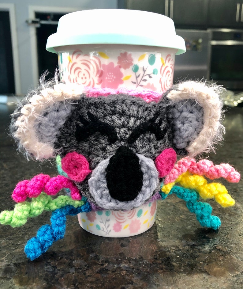 Puede incluir: Un calientavasos de ganchillo de koala con detalles de hilo arco&iacute;ris. El calientavasos es gris con detalles rosas y ojos negros. El calientavasos est&aacute; en una taza con estampado floral.