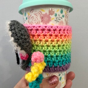 Puede incluir: Un calentador de taza de crochet arco&iacute;ris con un elefante de crochet gris y rosa adjunto. El calentador est&aacute; en una taza de cer&aacute;mica blanca con un patr&oacute;n floral y una tapa azul claro.