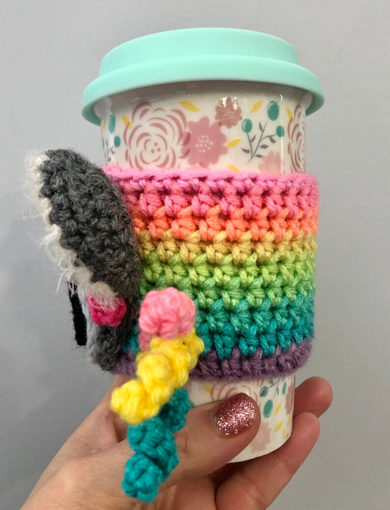 Puede incluir: Un calentador de taza de crochet arco&iacute;ris con un elefante de crochet gris y rosa adjunto. El calentador est&aacute; en una taza de cer&aacute;mica blanca con un patr&oacute;n floral y una tapa azul claro.