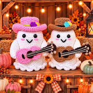 May include: Two crocheted ghost figures with black eyes and pink cheeks. One ghost wears a pink cowboy hat with flowers and holds a pink guitar. The other ghost wears a brown cowboy hat with flowers and holds a brown guitar. Both have white bodies.