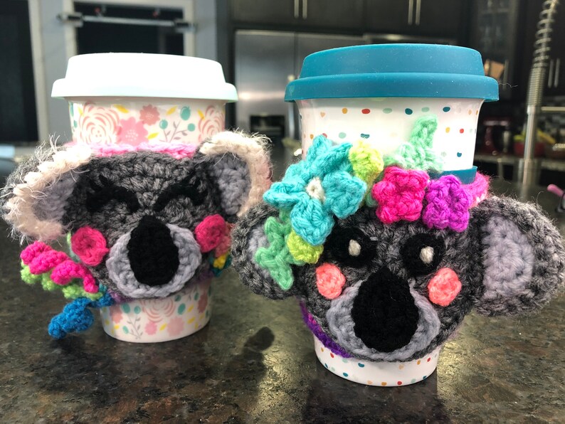 Puede incluir: Dos tazas de caf&eacute; con calientavasos de koala de ganchillo. Los calientavasos son grises con narices negras y mejillas rosadas. Una taza tiene un patr&oacute;n floral y la otra tiene lunares blancos. Ambos calientavasos tienen flores de colores.