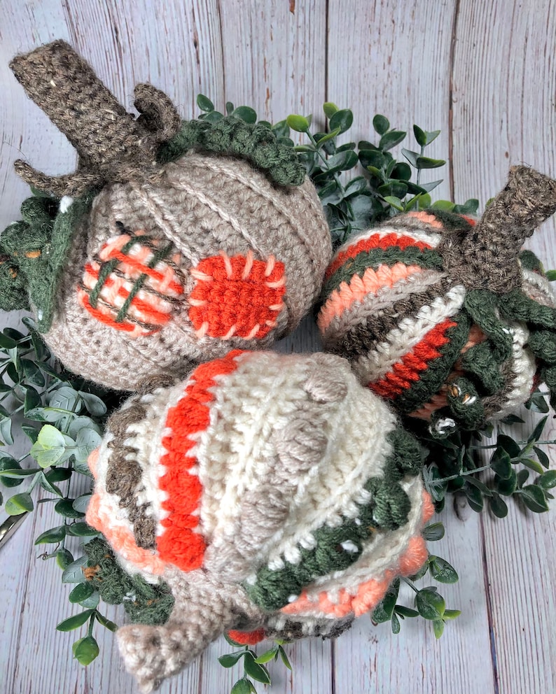 May include: Three handmade crochet pumpkins in shades of beige, orange, and green, arranged on a bed of greenery. Each pumpkin features a textured design, with a brown stem and green leaves. The pumpkins vary in size and pattern.