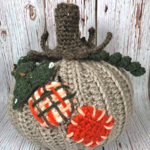 May include: A handmade crochet pumpkin in shades of gray, orange, and green. The pumpkin features a ribbed texture, a brown stem, and decorative patches in orange and plaid patterns. A fall-themed decorative item.