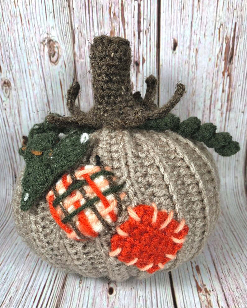 May include: A handmade crochet pumpkin in shades of gray, orange, and green. The pumpkin features a ribbed texture, a brown stem, and decorative patches in orange and plaid patterns. A fall-themed decorative item.