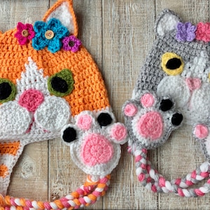 May include: Two crocheted cat hats with pink paw accents. One hat is orange with green eyes and the other is gray with yellow eyes. Both hats have white faces and pink noses.