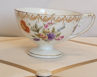 May include: A white porcelain teacup with a pedestal base, adorned with a floral pattern in shades of orange, pink, purple, and yellow. The rim features a gold filigree design, and the handle is also accented with gold.