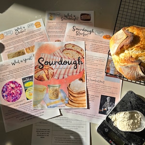 Könnte beinhalten: Ein frisch gebackenes Sauerteigbrot wird zusammen mit Lehrmaterialien zum Sauerteigbacken präsentiert. Das Material umfasst einen Studienführer, Rezeptkarten und Informationen zu Gluten und Hefe. Eine Digitalwaage mit Mehl ist ebenfalls vorhanden.