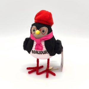 May include: A black and white plush bird wearing a red beret, pink scarf, and heart-shaped glasses. The bird is standing on red legs and has the word "Bonjour" embroidered on its chest.