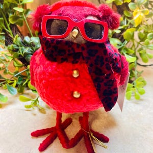 May include: A plush red bird figurine wearing red sunglasses and a black and red leopard print scarf. The bird has gold button accents and red feet. A decorative item.